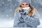 Woman wrapped in a cozy gray sweater and scarf, shielding themselves from falling snow while looking directly at the camera. The soft, fluffy hat and gloves add warmth to their winter attire against a snowy background.